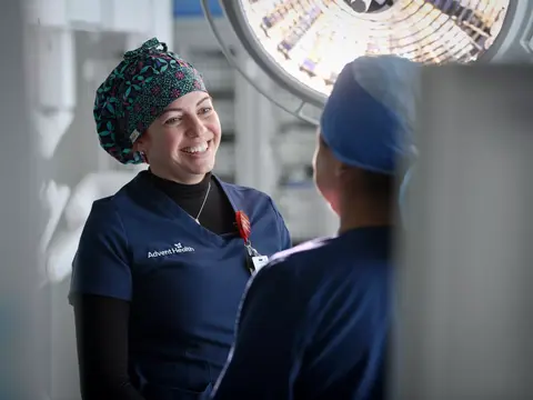 A smiling AdventHealth nurse in scrubs talks to another nurse in an operating room.