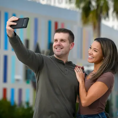 A smiling man and woman taking a selfie in front of a colorful building.