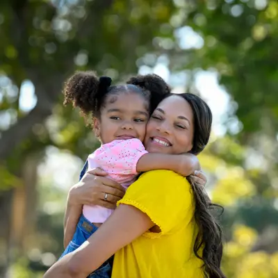 A smiling woman hugs her young daughter in a park, both looking happy and content.
