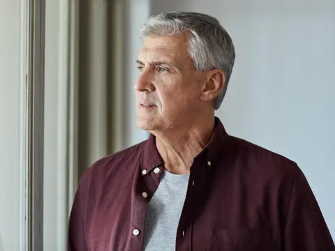 Older man standing indoors looking out a window.