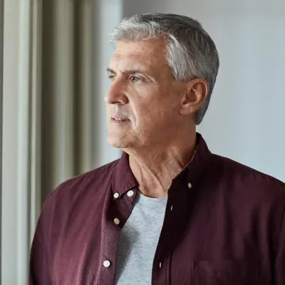 Older man standing indoors looking out a window.