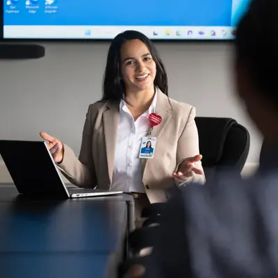 Health care professional in tan blazer engaging with colleagues during a meeting with laptop and monitor.