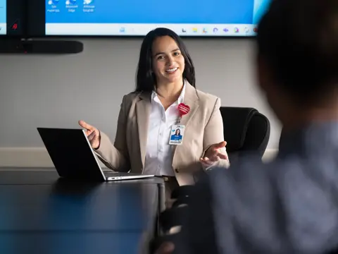 Health care professional in tan blazer engaging with colleagues during a meeting with laptop and monitor.