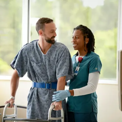A smiling man with a walker is supported by a nurse in a hospital room.