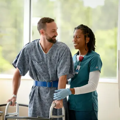 A smiling man with a walker is supported by a nurse in a hospital room.