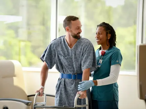 A smiling man with a walker is supported by a nurse in a hospital room.