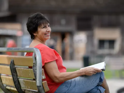 A smiling woman in a red top and blue jeans sits on a bench reading a book.