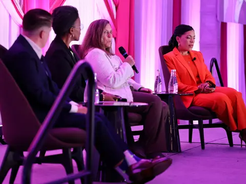 Four panelists seated on stage during a conference discussion, with one speaker addressing the audience with a microphone.