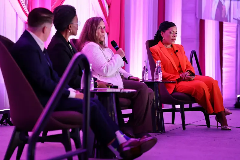 Four panelists seated on stage during a conference discussion, with one speaker addressing the audience with a microphone.