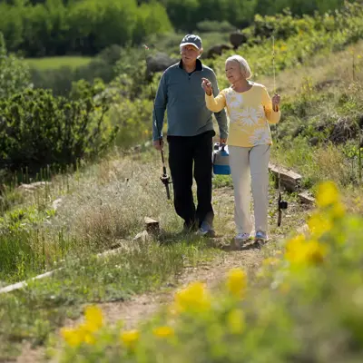 An elderly couple walking along a path by a lake, holding fishing rods and chatting.