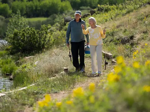 An elderly couple walking along a path by a lake, holding fishing rods and chatting.