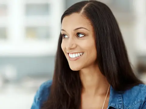 Smiling woman with long dark hair looks to the side, wearing a blue shirt, blurred indoor background.