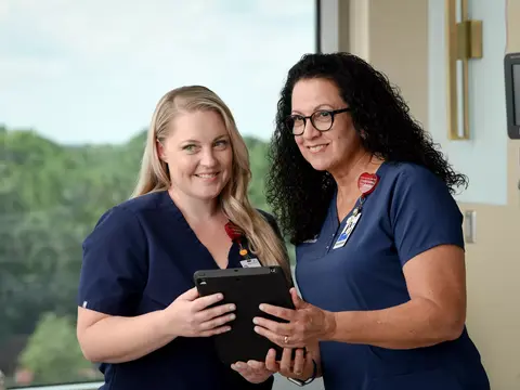 Two nurses in blue scrubs stand by a window, one holding a tablet, smiling.