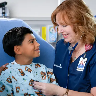 A smiling nurse with a stethoscope around her neck listens to a young boy's heart while his father watches.