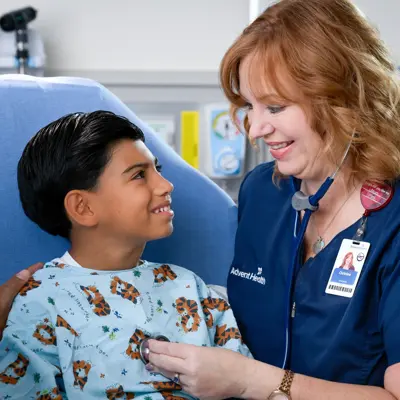A smiling nurse with a stethoscope around her neck listens to a young boy's heart while his father watches.