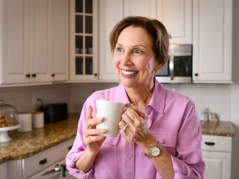 A smiling woman in a pink shirt holds a white mug in a modern kitchen.
