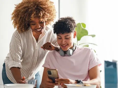 Two people smile while looking at a smartphone; one wears headphones around their neck.