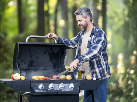 A middle-aged man in a blue plaid shirt grilling colorful vegetables on an outdoor grill in a natural wooded setting