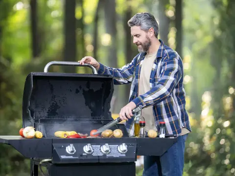 A middle-aged man in a blue plaid shirt grilling colorful vegetables on an outdoor grill in a natural wooded setting