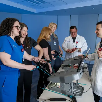 Six medical professionals in scrubs and white coats practicing laparoscopic surgical techniques on a training simulator in a blue-walled simulation room