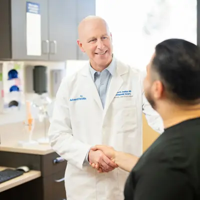 A smiling doctor in a white coat shakes hands with a patient in a medical office.