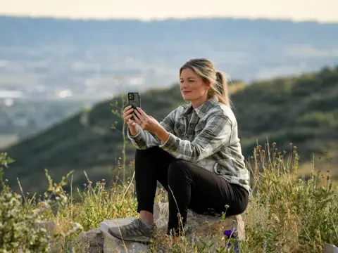 A woman sits on a rock, taking a photo with her smartphone, surrounded by nature.
