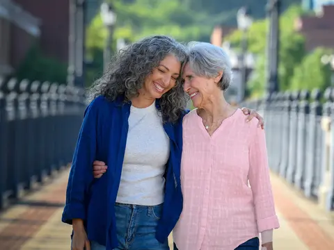 Two smiling women embracing outdoors on a sunny day, standing on a walkway with a metal railing and buildings in the background