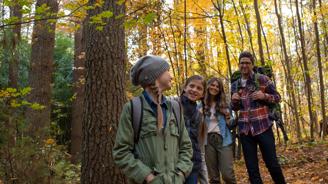 Group of hikers, including two adults and two children, smiling and exploring a forest with autumn leaves.