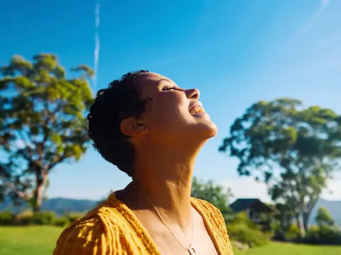 Woman with joyful expression looking up at the sky in a sunny outdoor setting with green grass and trees