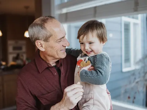 Older man in a maroon polo shirt smiling while holding a young child wearing a gray sweater and tan overalls with a heart-shaped cookie in a bright home kitchen