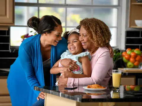 Three generations of women embrace in a cozy kitchen, sharing a joyful moment.