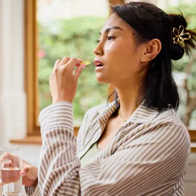 Woman taking a dietary supplement capsule with a glass of water while standing indoors near a window