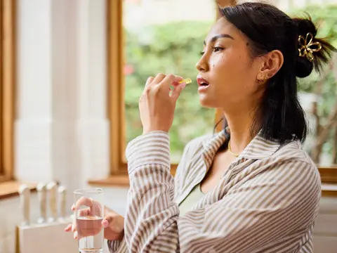 Woman taking a dietary supplement capsule with a glass of water while standing indoors near a window.