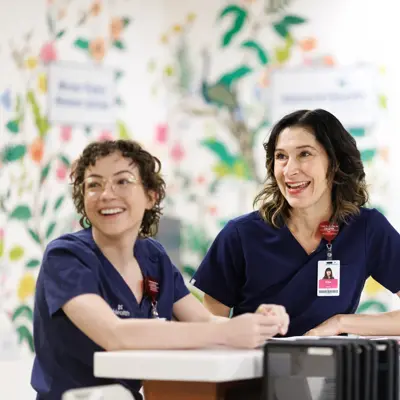 Two nurses in navy blue scrubs smiling and engaged in conversation with a person seated across from them at a table in a bright room with colorful floral wall art