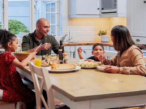 Family of four sharing a meal together at their kitchen table, with a father serving salad to his daughter while the mother and son smile