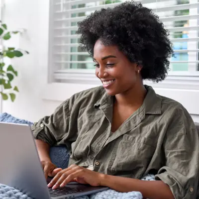 A smiling woman with curly hair is sitting on a couch using a laptop.