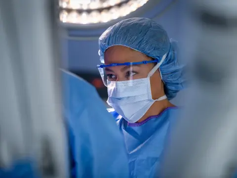 A focused surgeon in blue scrubs and protective gear stands in an operating room, ready for surgery.