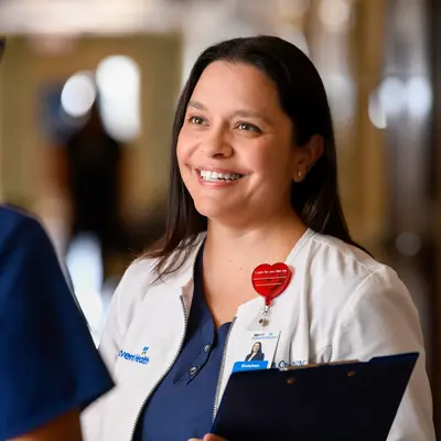 Two nurses in scrubs having a conversation in a hospital hallway, one wearing a white coat over blue scrubs and holding a clipboard, smiling warmly.