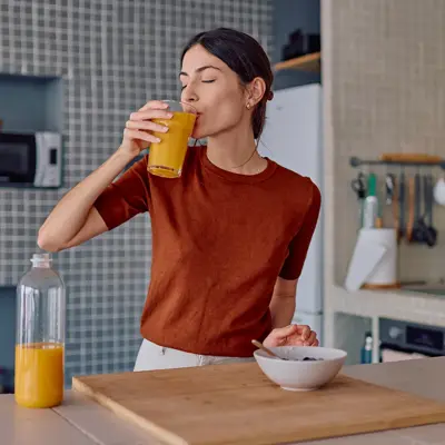 Woman in an orange shirt drinking a glass of orange juice in her kitchen with a bottle of juice and a bowl on the counter