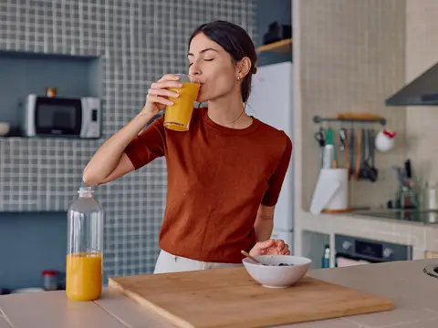 Woman in an orange shirt drinking a glass of orange juice in her kitchen with a bottle of juice and a bowl on the counter