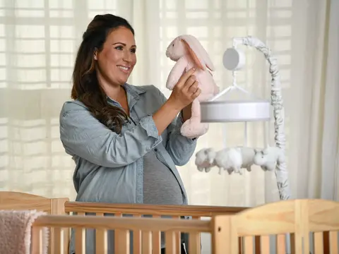 Pregnant woman holding a pink bunny toy in a nursery with crib and mobile.