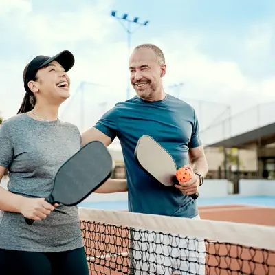 Two people holding pickleball paddles smile together across the net on an outdoor court.