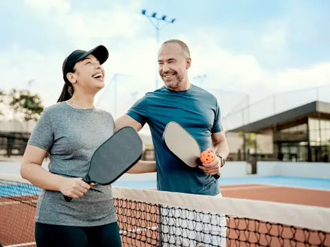 Two people holding pickleball paddles smile together across the net on an outdoor court.
