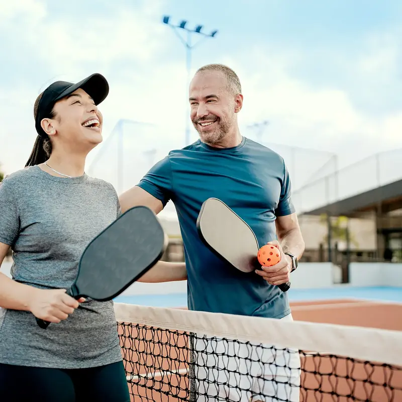 Two people holding pickleball paddles smile together across the net on an outdoor court.