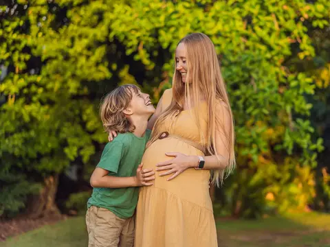 Pregnant woman and young boy smiling together outdoors, with the boy resting his hands on her belly.