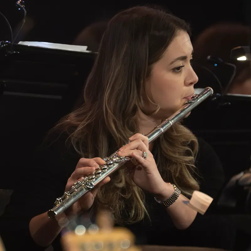 Erica Kopp performs with an orchestra, holding a silver flute beneath music stand lights.