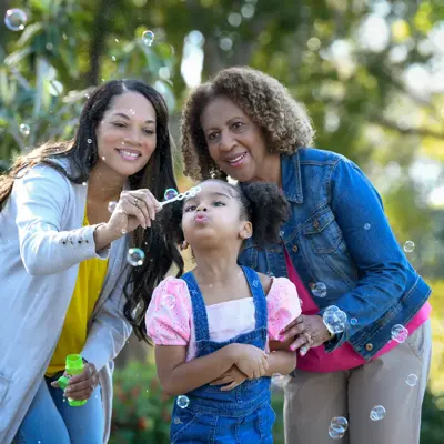 Three generations of women smiling and blowing bubbles in a park.