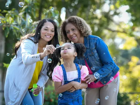 Three generations of women smiling and blowing bubbles in a park.