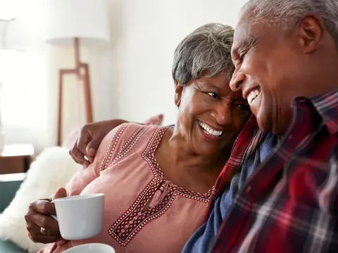 Two older adults sit close on a couch, smiling and holding mugs in a bright living room.