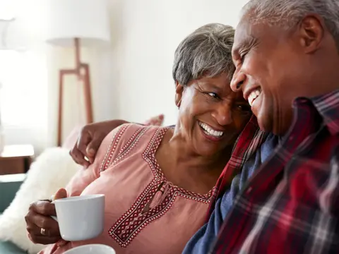 Two older adults sit close on a couch, smiling and holding mugs in a bright living room.